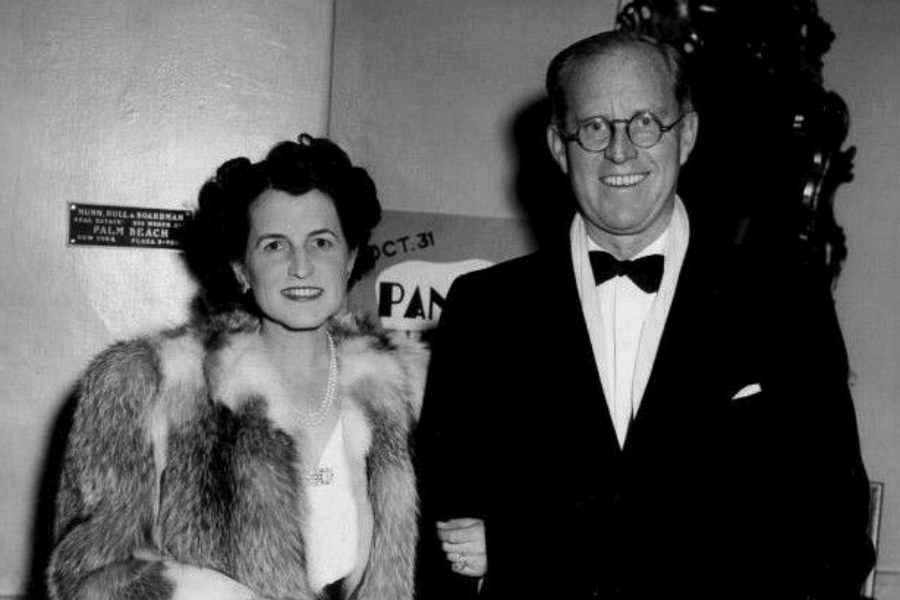 A black-and-white photograph of a formally dressed couple. The woman wears a luxurious fur coat over a gown, accessorized with pearls, and smiles warmly. The man stands beside her in a tuxedo with a bow tie and round glasses, also smiling, exuding elegance and sophistication in a glamorous setting.