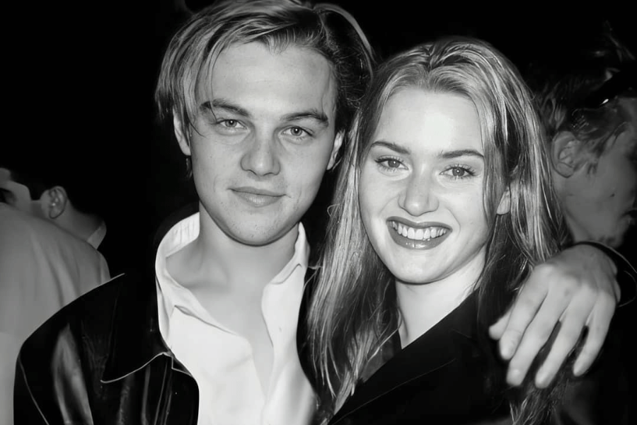 This black-and-white image features a young man and woman standing closely together, smiling warmly at the camera. The man has swept-back hair and wears a leather jacket over a partially unbuttoned shirt, while the woman has sleek hair and a radiant smile, dressed in a dark outfit. The background is blurred, suggesting a social event or gathering. 