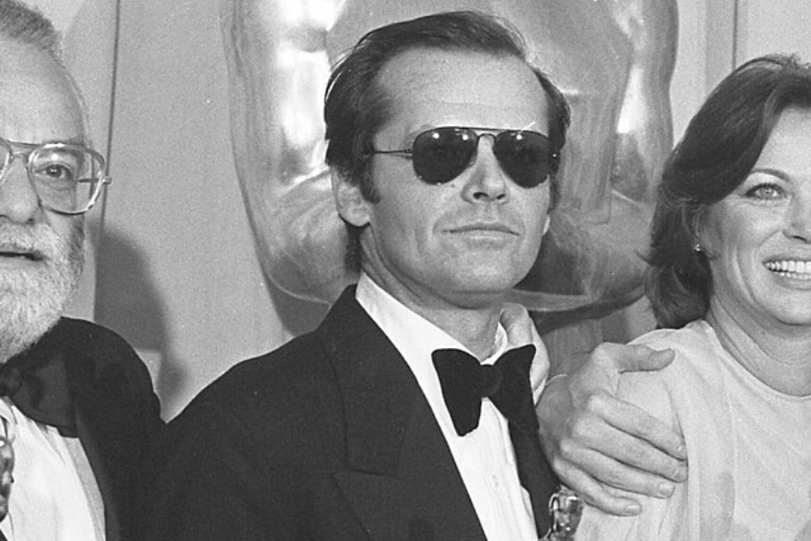 This black-and-white image shows a person wearing a tuxedo, bow tie, and dark aviator sunglasses, standing between two others, likely at a formal event. The background includes a large statue resembling an award, suggesting a prestigious ceremony.