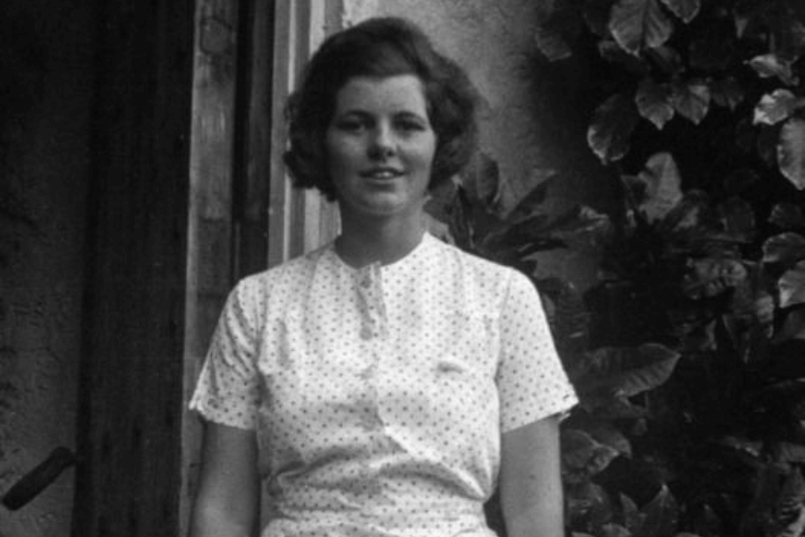 A black-and-white photograph of a young woman standing outdoors, wearing a short-sleeved, button-up dress with a subtle polka dot pattern. Her hair is styled in a soft, natural bob, and she has a calm expression, with leafy greenery visible in the background.