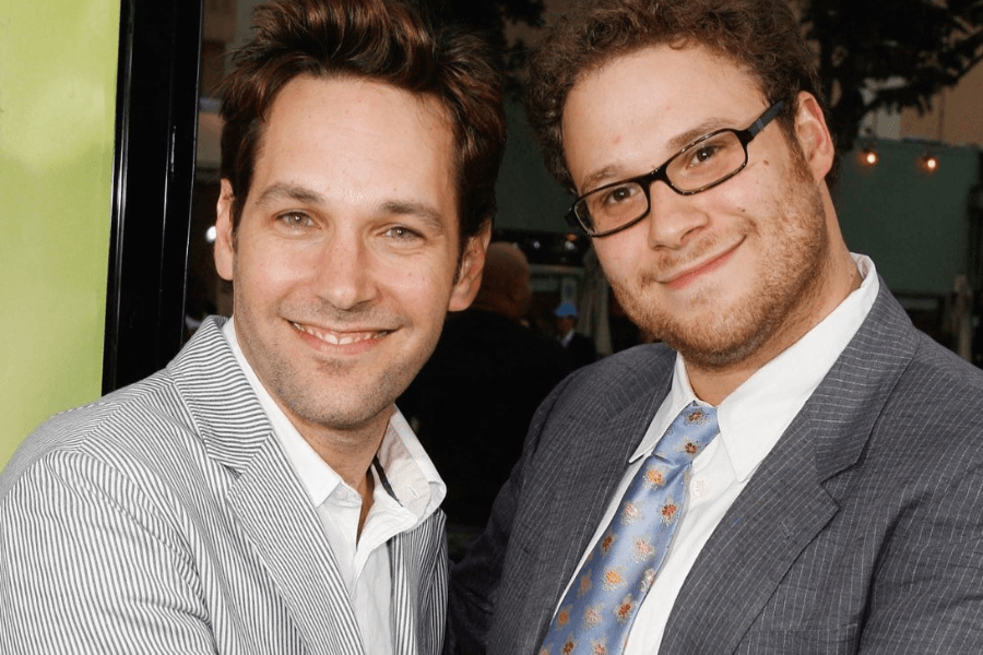 This image features two men smiling warmly for the camera in an outdoor setting, possibly at a film or media event. One man wears a light pinstripe blazer over a white shirt, exuding a casual yet polished look, while the other, sporting glasses, is dressed in a dark suit with a patterned tie. The background includes blurred trees and lights, adding to the festive atmosphere.