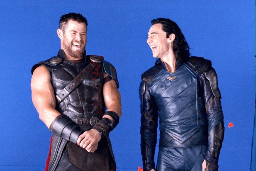 This image shows two men in elaborate costumes, laughing together on a set with a blue screen backdrop, indicating a behind-the-scenes moment during a film production. One man, wearing a muscular leather armor costume with straps, exudes a warrior-like appearance, while the other, dressed in sleek, dark armor with a mischievous look, adds contrast. Their genuine laughter suggests camaraderie and lightheartedness during filming. 