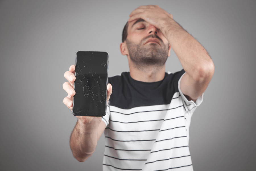  A man in a striped shirt holds up a smartphone with a cracked screen while covering his face with his other hand, appearing frustrated or upset. The background is a neutral gray.