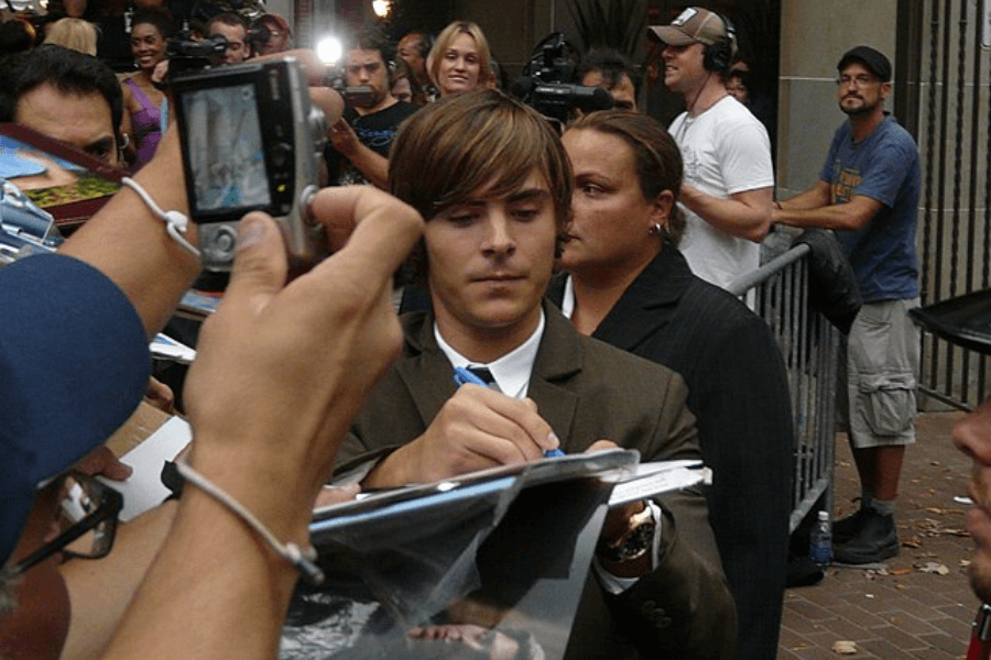 This image shows a person dressed in a suit and tie signing autographs for fans in a crowded outdoor setting. The scene is bustling with people taking photos, holding items to be signed, and a security person standing nearby to manage the event.