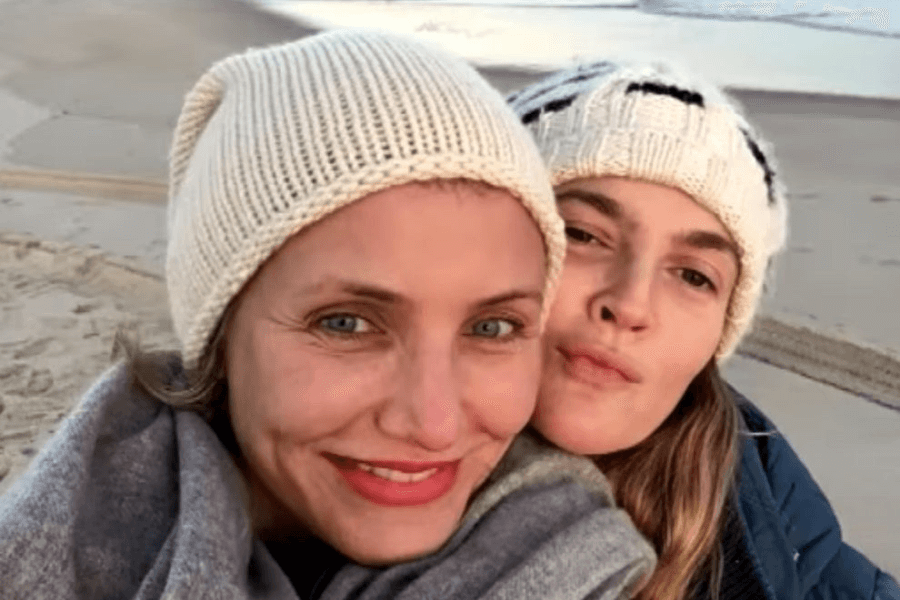 This image features two women bundled up in cozy winter attire, including knit beanies, as they pose for a close-up selfie on a beach. One smiles warmly, while the other playfully pouts. The sandy shoreline and water in the background add to the serene, outdoor setting.