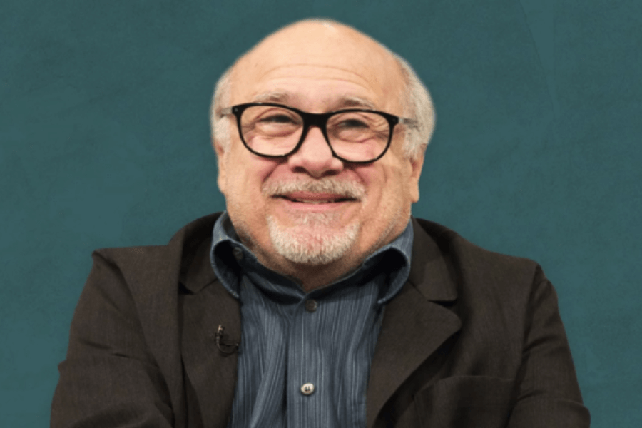 This image features a person with glasses, a short beard, and a warm smile, dressed in a dark blazer and a striped shirt. The background is a solid teal color, creating a simple and clean composition.