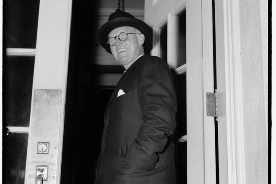 A black-and-white photograph of a man dressed in a suit, overcoat, and hat, standing by an open door. He smiles warmly as he glances back, with one hand casually in his pocket. The image exudes a sense of confidence and approachability in a candid moment.