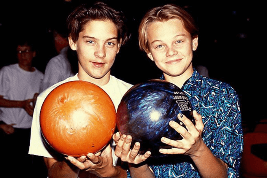 This image shows two young boys holding bowling balls, one orange and one blue, while posing for the camera in a bowling alley. The boy on the left wears a white shirt and has a relaxed expression, while the boy on the right, dressed in a colorful patterned shirt, smiles warmly. The background includes blurred figures and dim lighting typical of a bowling alley atmosphere.