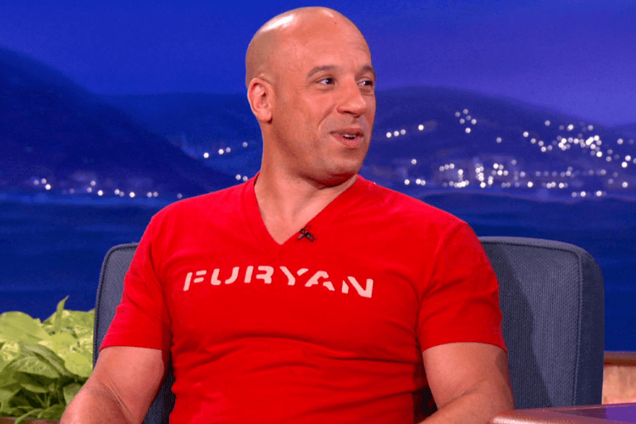 This image features a person seated on a talk show set, wearing a red t-shirt with the text "FURYAN" printed on it. The background displays a nighttime coastal scene with twinkling lights and dark hills in the distance.