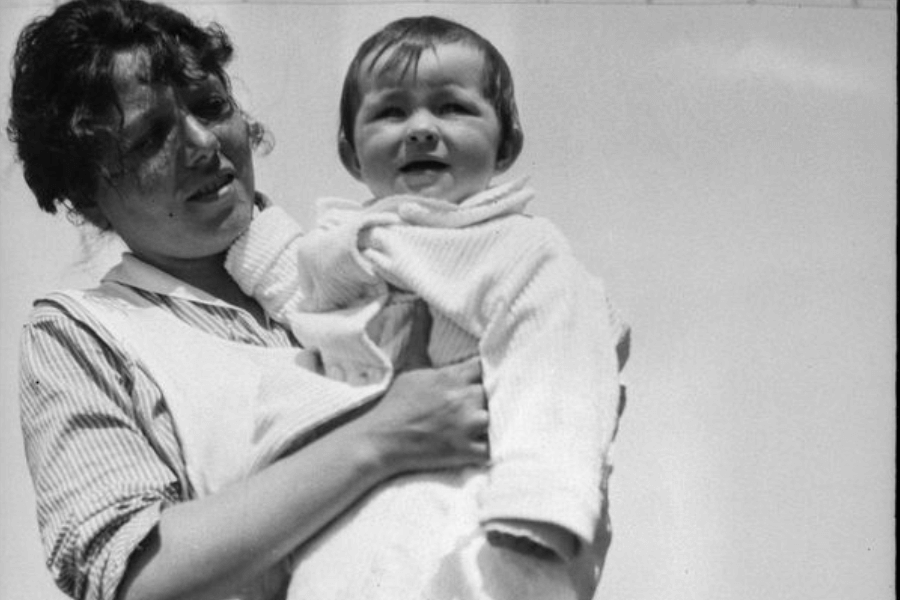 A black-and-white photograph of a woman holding a smiling baby in her arms against a bright, clear sky. The woman wears a striped blouse and apron, looking at the child with a soft, caring expression, while the baby is dressed in a cozy outfit and looks joyful.