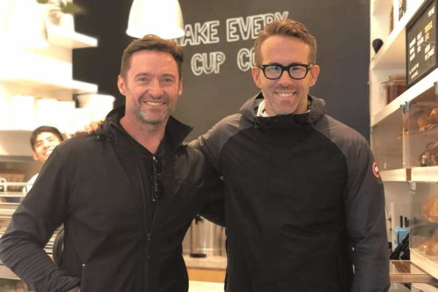 
This image shows two men standing side by side in a warmly lit café or casual dining setting. Both are smiling, with one wearing glasses and a black jacket with a logo on the sleeve, and the other dressed in a black zip-up top with sunglasses hanging from the collar. A chalkboard in the background displays part of a phrase, “MAKE EVERY CUP COUNT,” adding to the friendly and welcoming atmosphere. 