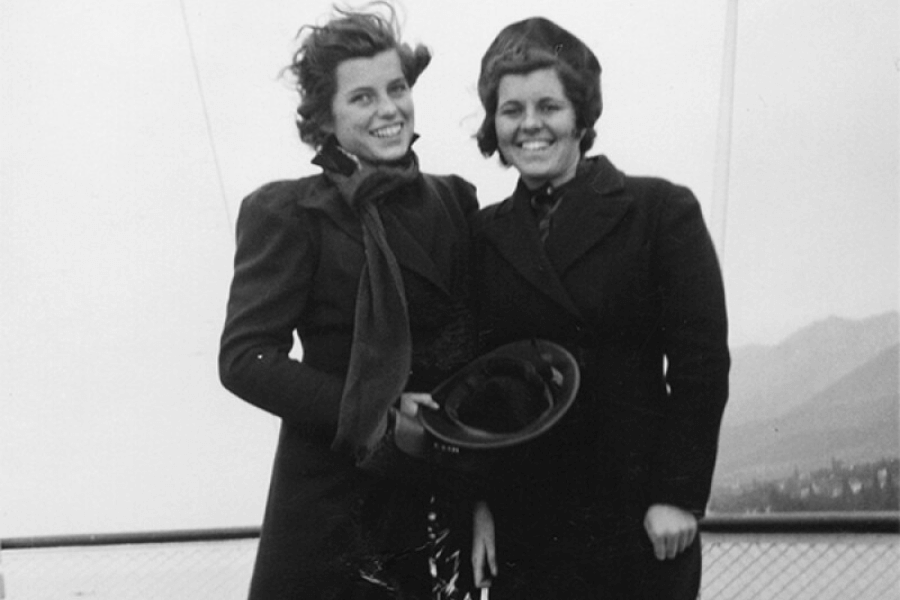 A black-and-white photograph of two women standing outdoors, smiling brightly despite the windy conditions, as evident from their windblown hair. Both are dressed in long coats and scarves, with one holding a hat in her hands. The background features faint mountains and an overcast sky, adding a sense of adventure and camaraderie to the scene.