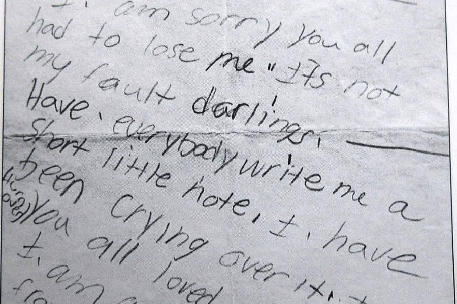 A close-up black-and-white image of a handwritten note with visible emotional content. The writing includes phrases like "I'm sorry you all had to lose me" and "it's not my fault, darlings," indicating it is a heartfelt message. The handwriting is informal, with a tone that appears personal and reflective.