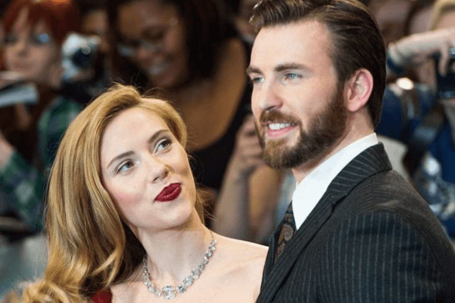 
This image shows a man and a woman posing at what appears to be a red carpet event, surrounded by a lively crowd of fans and photographers in the background. The woman, with wavy blonde hair, wears an elegant red dress and a sparkling necklace, looking at the man with a warm expression. The man, with neatly combed hair and a beard, is dressed in a striped suit and tie, smiling confidently. 