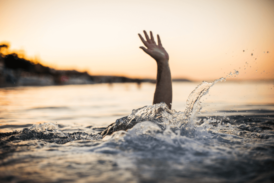 A single hand reaches above the water's surface as waves splash around, suggesting someone struggling in the water. The background features a calm sunset with warm orange hues over a blurred shoreline. 