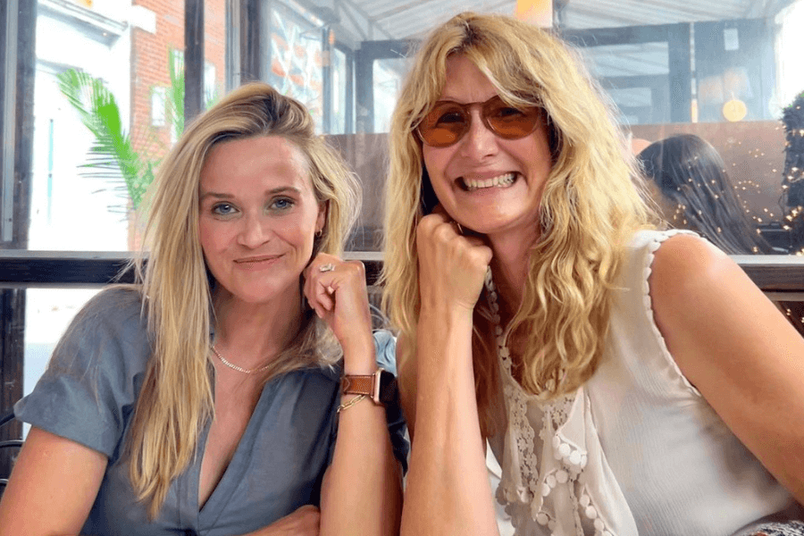 This image features two women sitting at an outdoor café or restaurant, smiling warmly at the camera. One has straight blonde hair and wears a blue blouse with a watch, while the other has wavy blonde hair, wears tinted glasses, and is dressed in a sleeveless white top. The setting includes a glass enclosure with a bright, relaxed atmosphere, suggesting a casual and friendly outing.