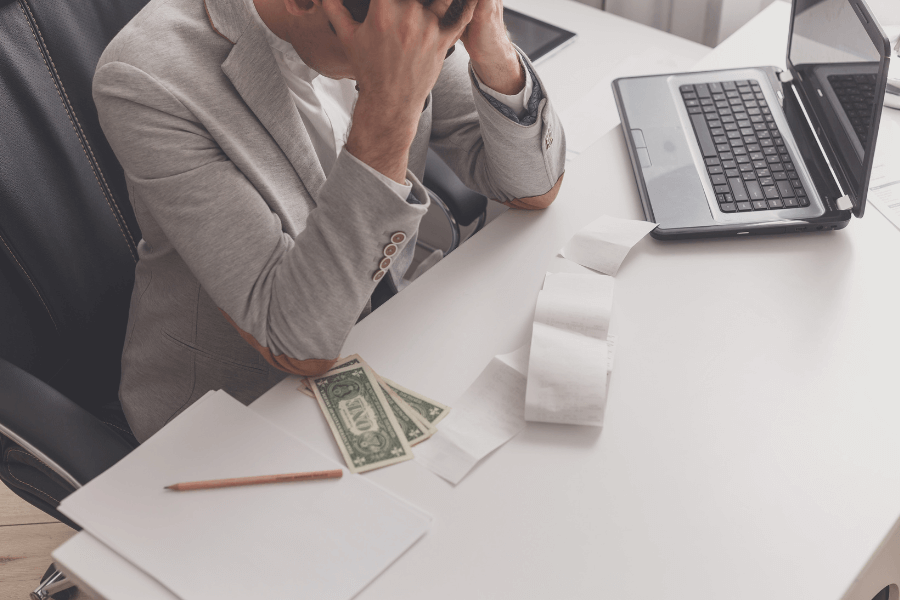 A man in a gray suit sits at a desk with his head in his hands, appearing stressed. On the desk are a laptop, receipts, a few dollar bills, and a notebook with a pencil, suggesting financial or work-related stress. 