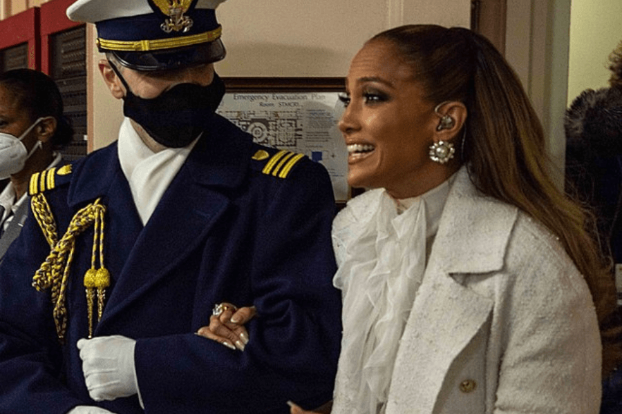 This image features a person dressed elegantly in a white outfit with a ruffled blouse and statement earrings, smiling and walking arm-in-arm with a uniformed officer. The setting appears to be indoors with other individuals in the background wearing face masks, indicating a formal or ceremonial occasion.