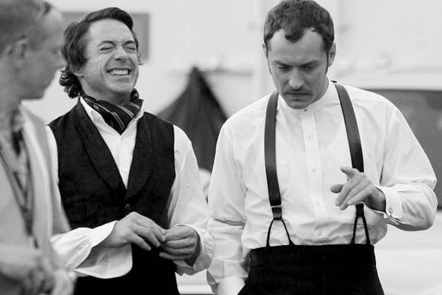 This black-and-white image captures two men dressed in period costumes, suggesting a behind-the-scenes moment during a film production. One man is smiling broadly, wearing a vest and scarf, while the other has a focused expression, dressed in a shirt with suspenders, gesturing as if discussing or rehearsing a scene. A third person in the foreground appears to be part of the crew. The casual and candid nature of the photo conveys a lighthearted yet professional atmosphere.