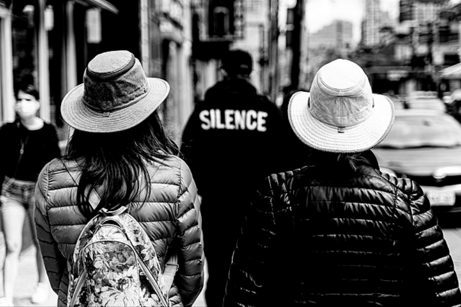 A black-and-white street photograph featuring two people wearing wide-brimmed hats and quilted jackets, walking away from the camera. One carries a floral-patterned backpack. In the background, a figure wearing a jacket with the word "SILENCE" printed on the back stands amidst an urban setting with buildings and cars, creating a contrast between motion and stillness.