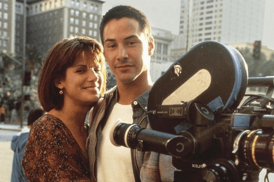 This image shows a man and a woman standing closely together, smiling near a professional film camera on a sunny day. The woman has a bob haircut and wears a patterned top, while the man, with short hair, wears a white shirt and light jacket. The backdrop includes urban architecture, suggesting they are on a movie set or location shoot. 