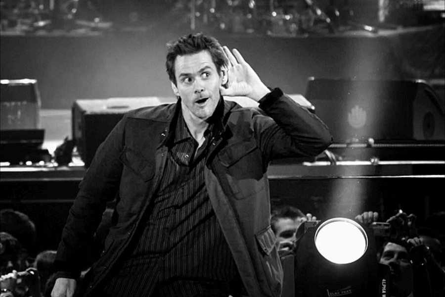 This black-and-white image shows a person on stage in a dynamic pose, leaning slightly forward and holding one hand to their ear with an expressive facial expression. The background features stage equipment and a crowd of spectators, suggesting a lively performance or event.