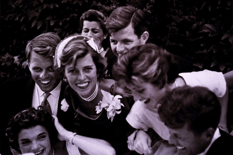 A candid, sepia-toned photograph of a joyous group of people gathered closely together, smiling and laughing. The central woman, wearing pearl jewelry and a floral corsage, exudes elegance and happiness. The scene is vibrant and lively, capturing a moment of celebration or camaraderie in a lush outdoor setting.