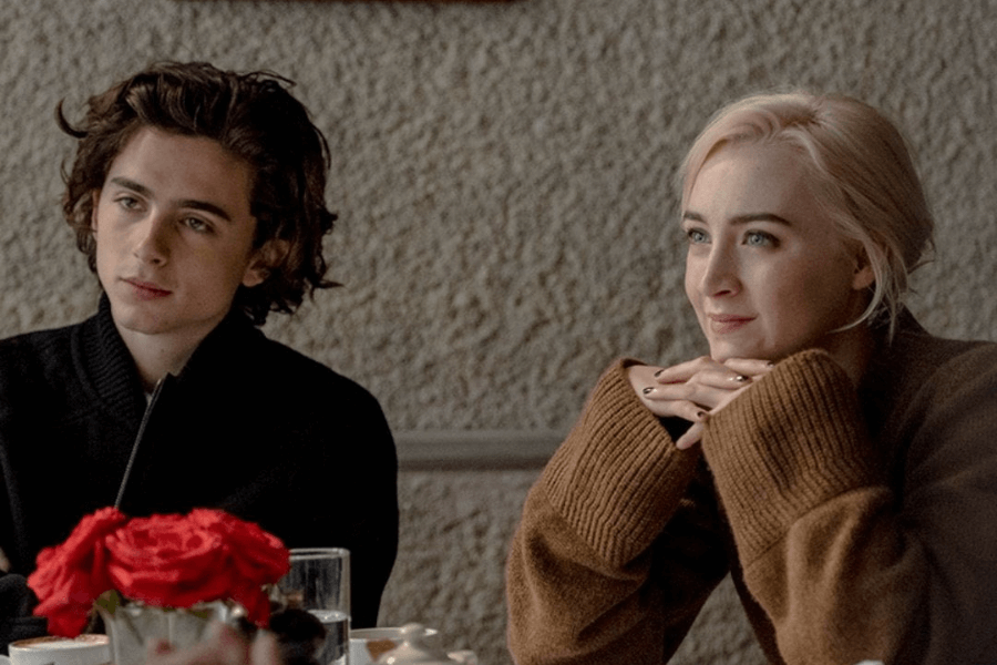 
This image features a young man and woman seated at a table in a cozy indoor setting. The man has dark, wavy hair and wears a dark sweater, looking contemplative, while the woman, with blonde hair and wearing a brown sweater, leans forward with her hands under her chin, smiling softly. A vase of red roses and glasses of water on the table add to the intimate, relaxed atmosphere. 