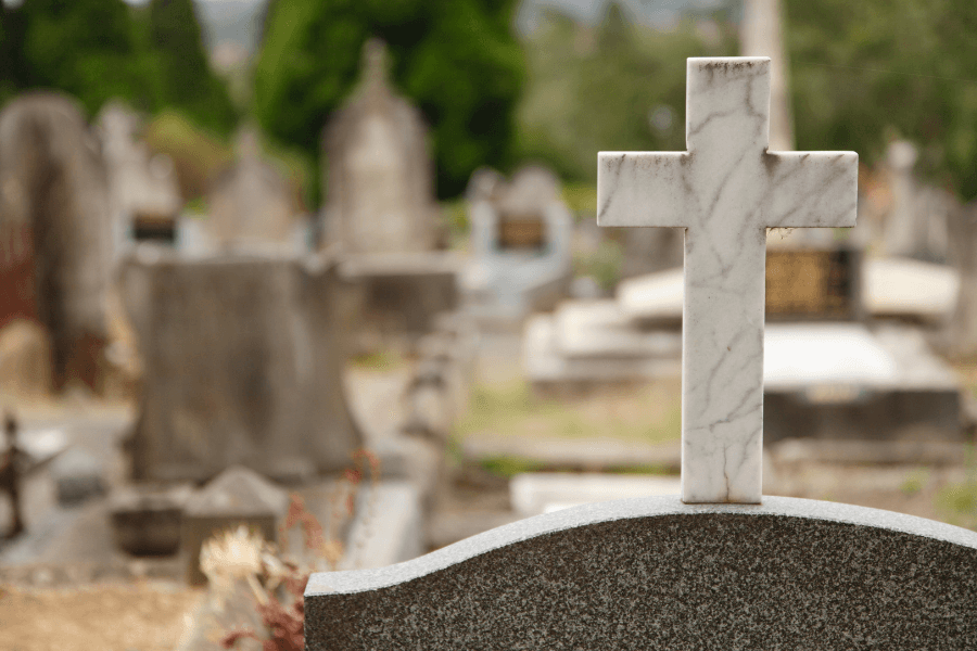  A white marble cross stands atop a gravestone in a cemetery, surrounded by other tombstones and blurred greenery in the background. The scene conveys a sense of solemnity and remembrance.