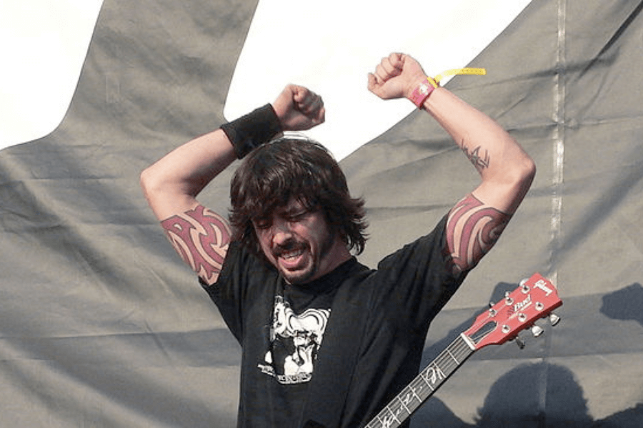 This image depicts a musician with long dark hair and tattoos on both arms, raising their fists in an energetic pose while holding a red electric guitar. They are wearing a black t-shirt and performing outdoors, with a large stage backdrop in the background.