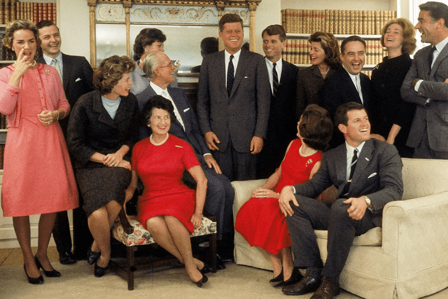 A vibrant color photograph capturing a lively group of people in a formal yet relaxed setting, laughing and chatting together. The group is dressed in elegant mid-20th-century attire, with some seated on a sofa and chairs, while others stand behind them. The backdrop includes bookshelves and a mirror, adding a refined, familial ambiance to the joyous scene.