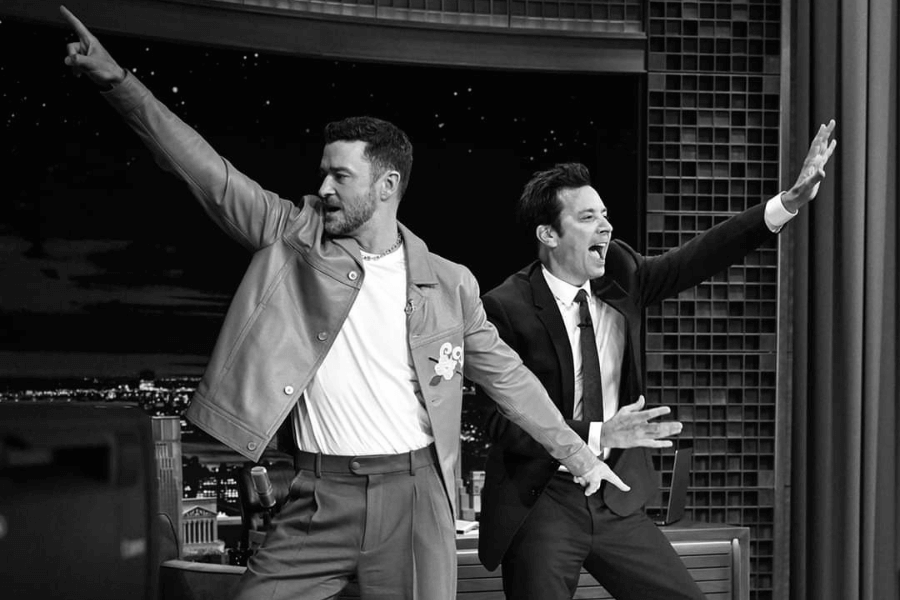 
This black-and-white image captures two men enthusiastically posing on a stage or television set. One wears a casual outfit with a jacket and white T-shirt, pointing upward dramatically, while the other, dressed in a suit, matches the energy with a wide grin and an outstretched arm. The lively, celebratory mood is enhanced by the nighttime city backdrop and studio setting.