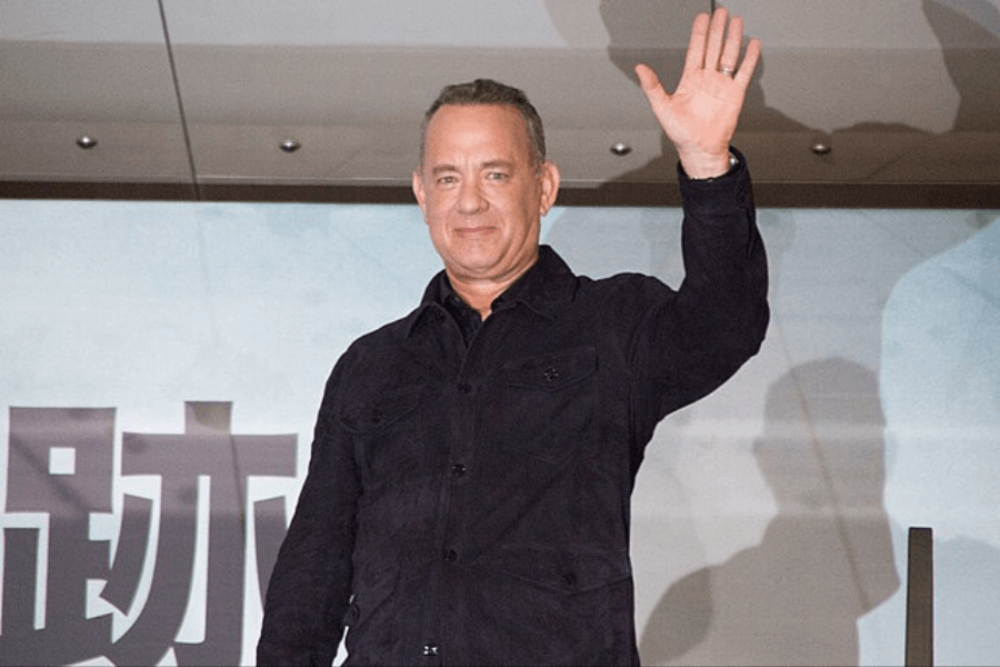 This image shows a person waving with a friendly smile while wearing a dark jacket, standing in front of a backdrop featuring large characters in a bold font. The setting appears to be an indoor event or promotional appearance.