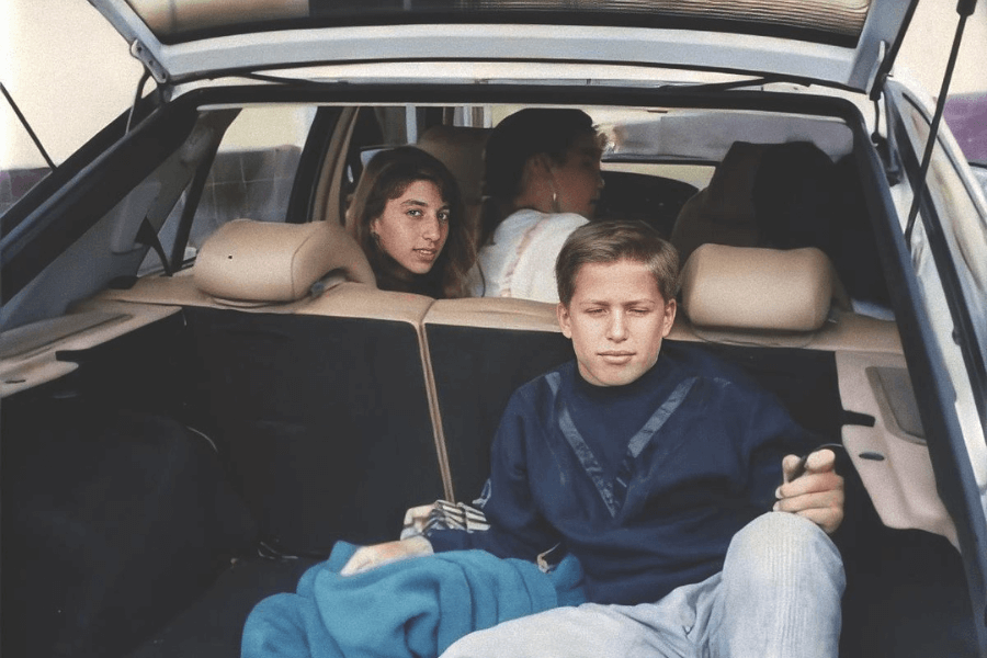 This image shows two children sitting in the open trunk of a car, with a third person in the front seat partially visible. The boy in the foreground is wearing a dark sweatshirt and appears relaxed, while the girl behind him looks toward the camera with a calm expression. The car’s interior and surroundings suggest a casual, everyday moment, possibly during a trip or outing.