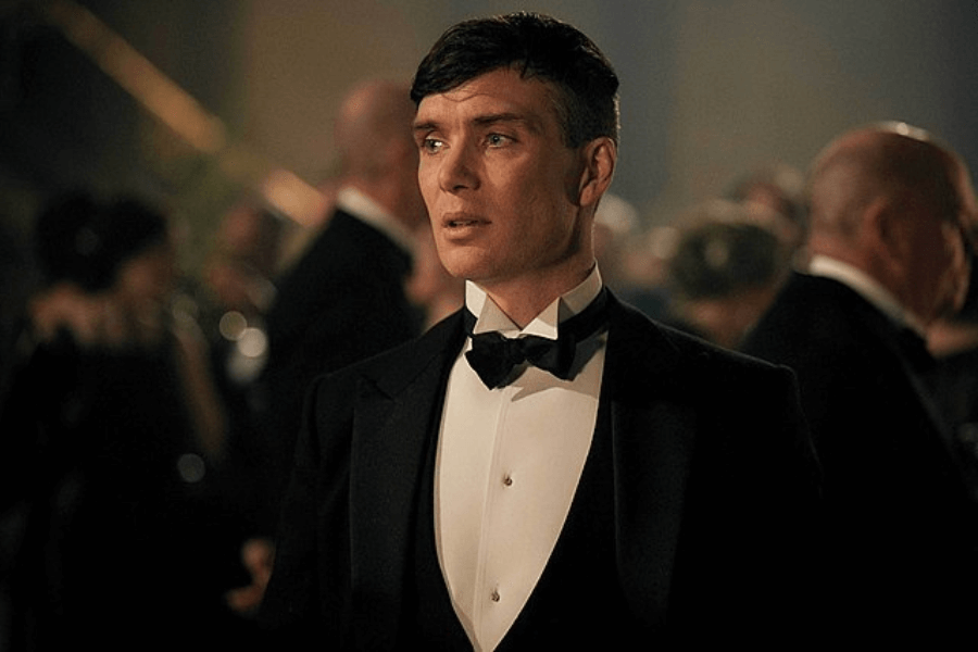 This image depicts a person dressed in an elegant black tuxedo with a bow tie, standing in a formal setting with a blurred background of other individuals in evening attire. The lighting and setting suggest a glamorous event, such as a gala or period-themed gathering.