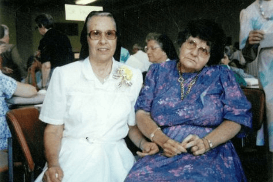 A color photograph of two elderly women seated next to each other at an indoor gathering. One woman is dressed in a white outfit with a corsage pinned to her chest, while the other wears a purple floral dress with glasses and a gold necklace. The setting includes other people and tables in the background, suggesting a social event or celebration.