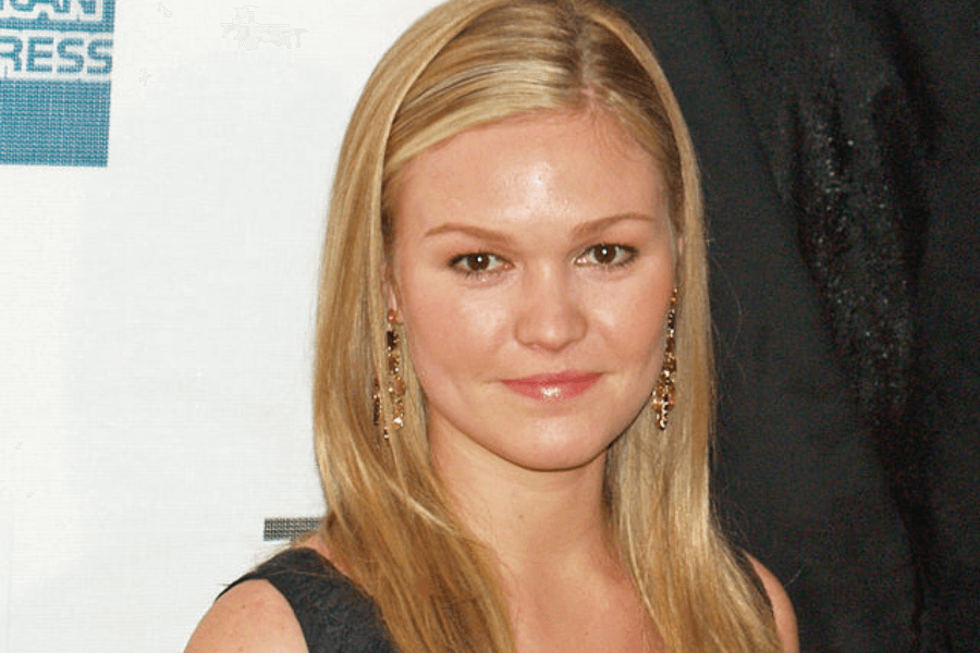 This image shows a person with long blonde hair wearing a dark sleeveless outfit and dangling earrings, posing against a white backdrop featuring text and logos, suggesting a formal event or premiere. The expression is poised and confident, fitting the occasion.