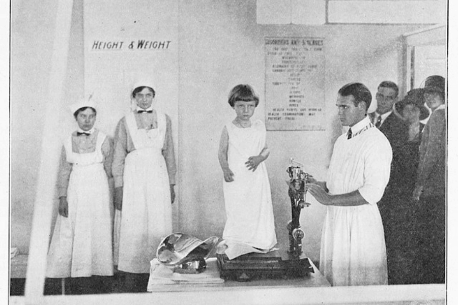 A vintage black-and-white photograph showing a young child standing on a scale during a height and weight measurement. The child is draped in a plain garment, while a man in a white coat adjusts the scale. In the background, three nurses in traditional uniforms and hats stand near a sign that reads "Height & Weight," and several onlookers are visible, observing the scene.