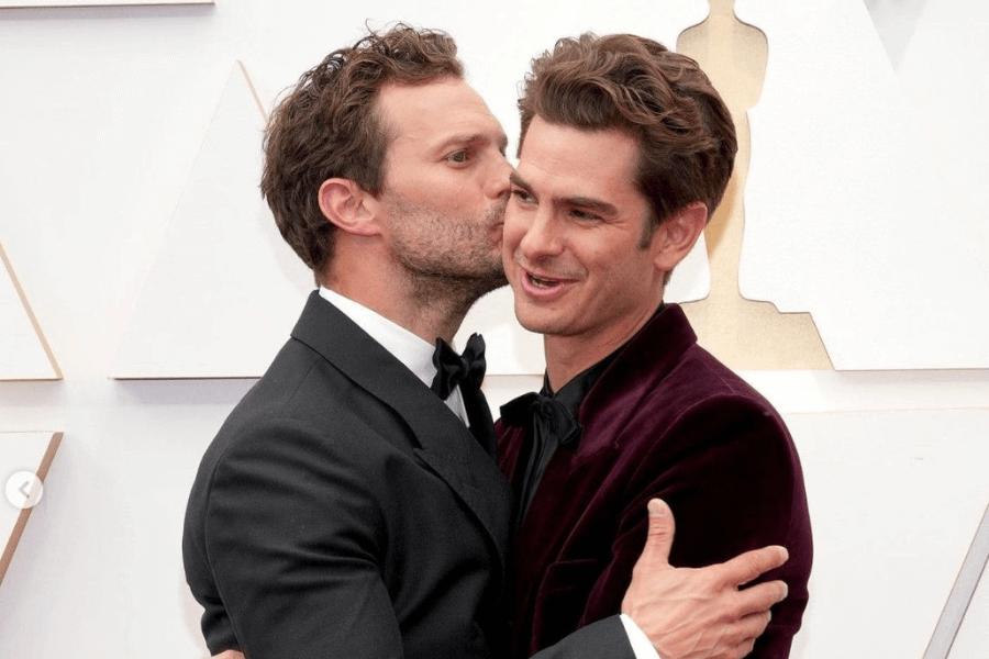 
This image captures a warm, playful moment between two men at a formal event, likely an awards ceremony. One man in a classic black tuxedo is kissing the other, who is wearing a velvet burgundy suit, on the cheek while they embrace. The background features geometric designs and a golden Oscar-like figure, emphasizing the glamorous setting