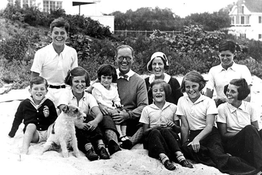 A black-and-white family photo taken outdoors on a sandy area with a backdrop of greenery and houses. The group consists of several children and adults, all smiling and seated closely together, with a small dog in the foreground. The image captures a joyful, relaxed moment, radiating familial warmth and connection.