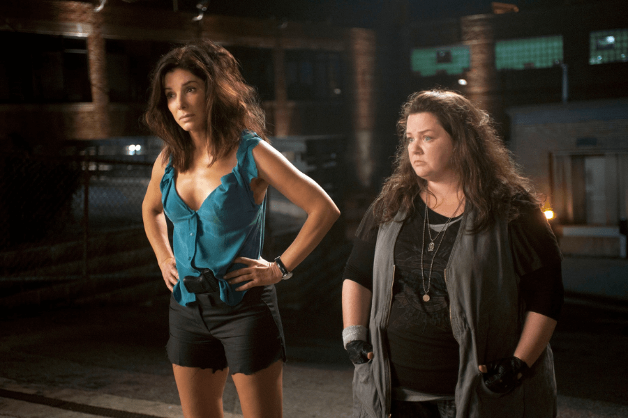 
This image shows two women standing outdoors at night in what appears to be an urban or industrial setting. One wears a sleeveless teal blouse and black shorts with her hands on her hips, exuding confidence or frustration, while the other wears a layered outfit with gloves, looking serious or contemplative. The background features brick buildings, chain-link fencing, and glowing windows, adding a gritty, cinematic tone. 