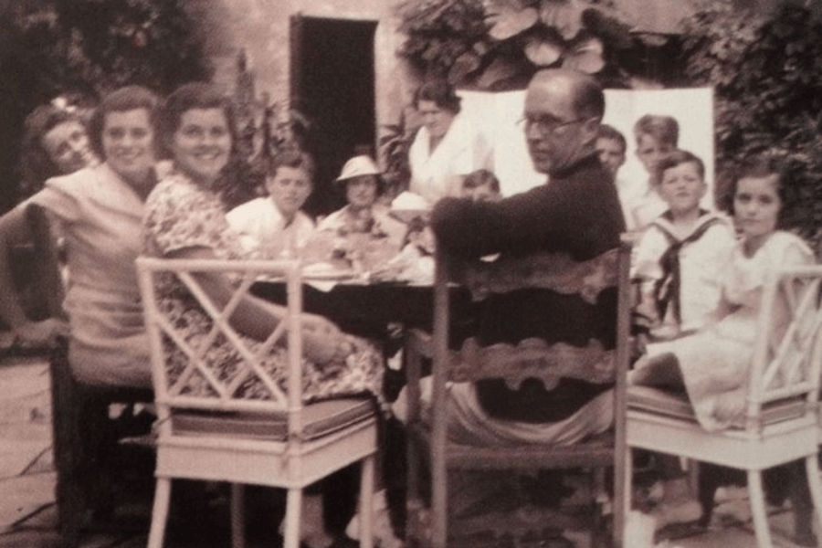 A sepia-toned photograph of a large family seated around a table in an outdoor courtyard setting. The group includes men, women, and children, all dressed in casual or semi-formal attire, engaging in conversation and sharing a meal. The background features lush plants and a rustic building, creating a warm and intimate atmosphere.