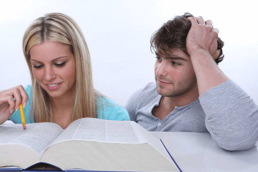 A young woman with long blonde hair smiles while studying a large open book, holding a pencil in her hand. Beside her, a young man with brown hair and a gray shirt leans on his hand, looking at her with a thoughtful expression. 