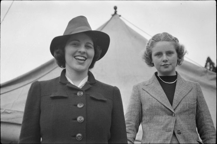 A black-and-white photograph of two women outdoors, with a tent visible in the background. One woman, wearing a wide-brimmed hat and a buttoned coat, smiles brightly, while the other, dressed in a plaid blazer and pearl necklace, looks composed and serious. The image captures contrasting expressions against a simple, open-air setting.