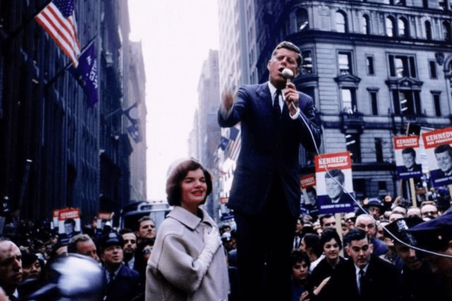 A vibrant color photograph of a political rally in a bustling city street, featuring a man in a suit speaking passionately into a microphone on a raised platform. A woman stands beside him, elegantly dressed in a coat and gloves, smiling at the crowd. The background is filled with supporters holding signs, American flags, and towering buildings, capturing the energy and excitement of the event.