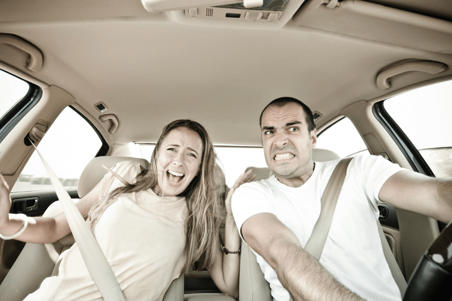 A man with a tense expression grips the steering wheel while driving, and a woman in the passenger seat appears panicked, screaming with her hands raised. Both are wearing seat belts inside a car, with visible tension and stress on their faces.