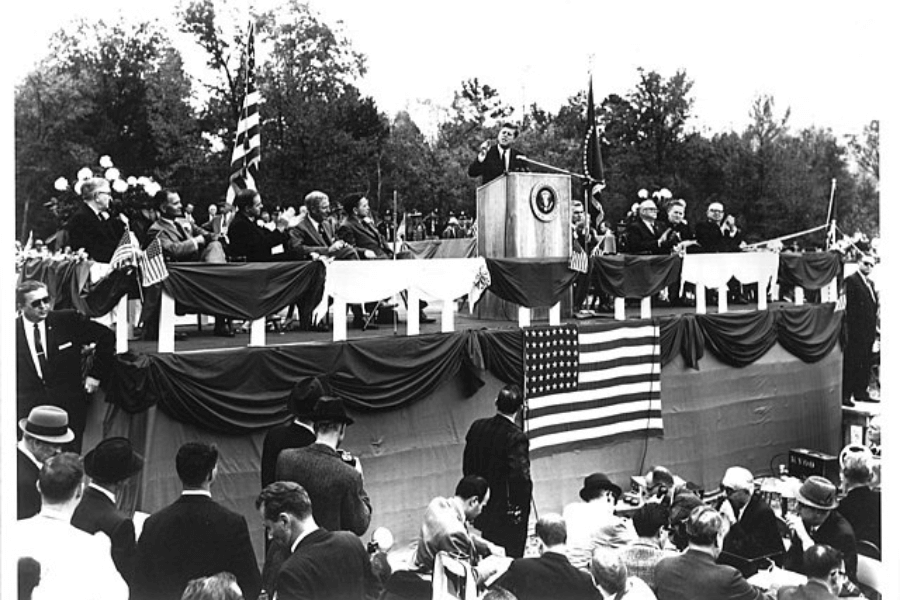 A black-and-white photograph of a formal outdoor event featuring a speaker at a podium decorated with the presidential seal. The stage is adorned with draped American flags, with dignitaries seated behind the podium. A large crowd gathers in the foreground, attentively watching the proceedings, set against a backdrop of trees, symbolizing a significant public or political occasion.