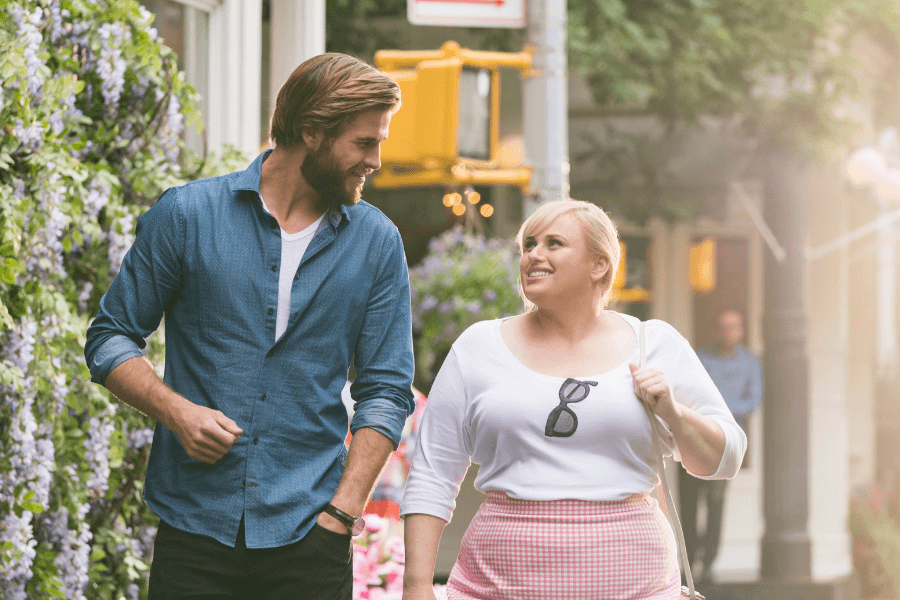 
This image features a man and a woman walking together in a sunny, charming urban street lined with greenery and flowers. The man, wearing a blue button-up shirt and a white T-shirt, smiles warmly as he talks to the woman, who is dressed in a white top, pink gingham skirt, and sunglasses hanging at her neckline. The soft lighting and casual atmosphere suggest a friendly and lighthearted moment.