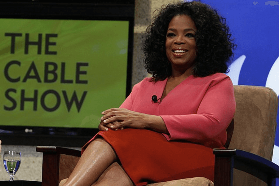 This image features a person seated on stage, wearing a pink top and an orange skirt, smiling warmly while participating in a discussion. A microphone is clipped to their outfit, and a backdrop displays the words "THE CABLE SHOW," suggesting a media or television-related event.