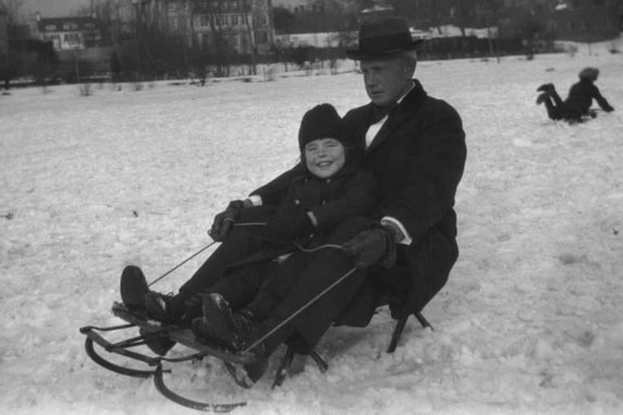 A black-and-white photograph capturing a playful winter scene of an adult and a child sledding together on snow. The child, bundled in warm winter clothing, grins with excitement, while the man in a coat and hat appears focused on guiding the sled. In the background, another person is seen sliding or playing in the snowy field, with buildings visible on the horizon.